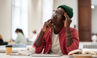 First-year college student looking confused while sitting at a desk with books and a laptop, representing the overwhelm many students feel before classes begin.